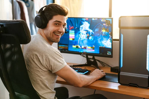 幸せなブルネット男でヘッドフォン再生ビデオゲームオン彼のコンピュータ座っている間に自宅 — ストック写真