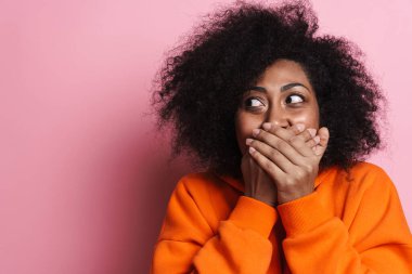 Neşeli Afro-Amerikalı kadın ağzını kapatıp pembe arka planda tek başına bir kenara bakıyor.