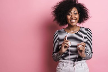 Neşeli Afrikalı Amerikalı kız pembe arka planda izole edilmiş kulaklıklarla poz verirken gülümsüyor.