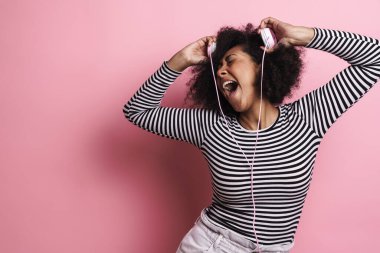 Neşeli Afrikalı Amerikalı kız pembe arka planda izole edilmiş kulaklıklarla dalga geçiyor.