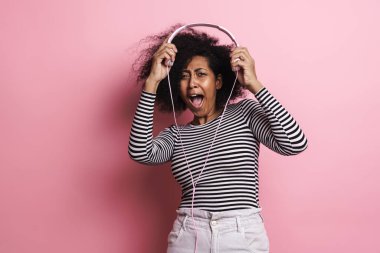 Neşeli Afrikalı Amerikalı kız pembe arka planda izole edilmiş kulaklıklarla dalga geçiyor.