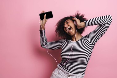 Neşeli, genç ve çekici Afrikalı bir kadın pembe arka planda izole edilmiş kulaklıklarla müzik dinliyor, dans ediyor.