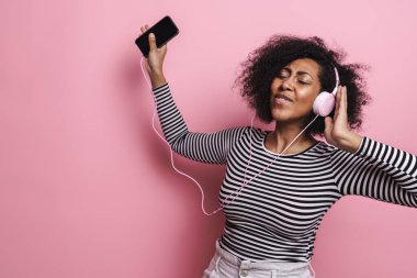 Neşeli, genç ve çekici Afrikalı bir kadın pembe arka planda izole edilmiş kulaklıklarla müzik dinliyor, dans ediyor.