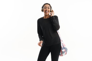 Beyaz arka planda izole edilmiş spor çantasıyla poz veren kulaklıklı, gülümseyen Afrikalı Amerikalı kadın.