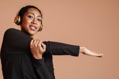 Yüzünde kulaklıkla gülümseyen Afrikalı Amerikalı kadın bej arka planda tek başına çalışırken egzersiz yapıyor.