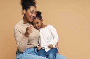 Mutlu siyah anne ve kız kucaklaşırken bej arka planda izole edilmiş kamerayı işaret ediyorlar.
