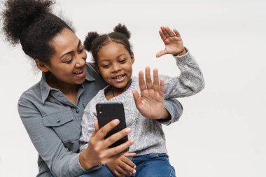 Beyaz duvarın üzerinden izole edilmiş cep telefonu kullanan Afrikalı anne ve kız el sallıyor.
