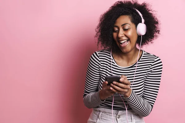 楽しいですアフリカ系アメリカ人の女の子リッスン音楽とともに携帯電話とヘッドフォン隔離されたピンクの背景 — ストック写真