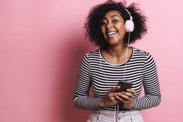楽しいですアフリカ系アメリカ人の女の子リッスン音楽とともに携帯電話とヘッドフォン隔離されたピンクの背景 — ストック写真