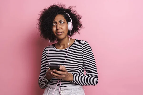 アフリカ系アメリカ人の女の子がピンクの背景に隔離された携帯電話やヘッドフォンで音楽を聞いて混乱 — ストック写真