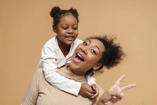 Happy black mother and daughter laughing while showing peace sign isolated over beige background