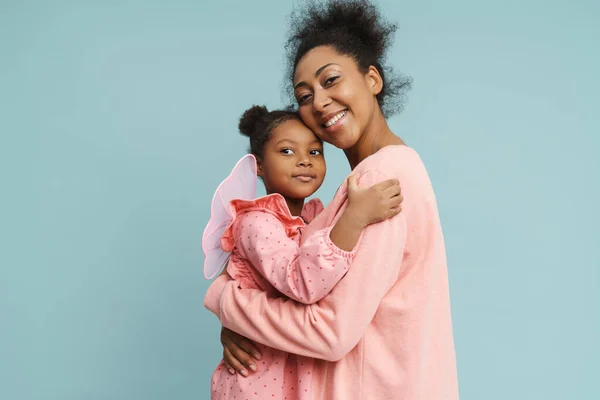 Happy black mother and daughter with butterfly wings hugging and smiling isolated over blue background