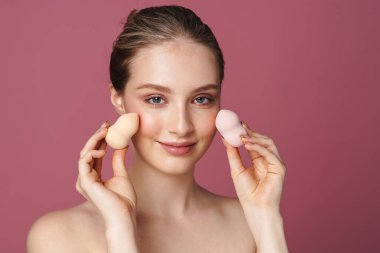 Pembe arka planda izole edilmiş kozmetik süngerlerle poz veren çekici genç bir kadının güzel portresini kapat.