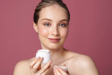Çekici genç bir kadının pembe arka planda izole edilmiş yüz kremi kavanozunu gösteren portresini kapat.