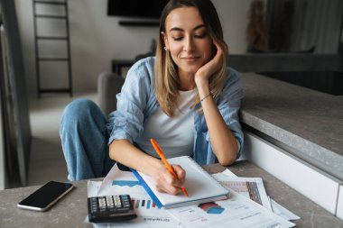 Evinde doküman ve hesap makinesiyle masada çalışan gülümseyen genç kadın.