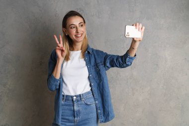 Gri bir arka planda izole edilmiş akıllı telefondan selfie çeken çekici bir kadının portresi.