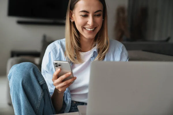 ラップトップコンピュータで家に座っている間 携帯電話を使用して幸せな若い女性 — ストック写真