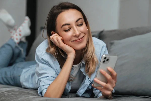 Χαμογελώντας Νεαρή Γυναίκα Χρησιμοποιώντας Κινητό Τηλέφωνο Ενώ Χαλαρώνοντας Έναν Καναπέ — Φωτογραφία Αρχείου