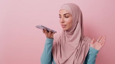 Güzel bir Arap kadın, stüdyodaki pembe bir duvarın üzerinde izole bir şekilde duran bir sesli mesaj kaydederken akıllı telefonunu kullanıyor.