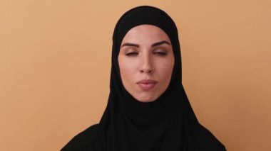 Siyah başörtüsü takan ciddi bir Arap kadının yakın plan görüntüsü, stüdyodaki bej bir duvarın üzerinde izole bir şekilde dururken olumsuz yanıt verirken başını sallıyor.