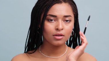 Güzel bir Afrikalı Amerikalı kadın, kaş fırçasıyla kaşlarını düzeltiyor. Stüdyonun gri duvarında tek başına duruyor.