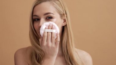 Çekici sarışın bir kadın, stüdyodaki bej bir duvarın üzerinde tek başına dururken yüzüne pudra fırçası sürüyor.