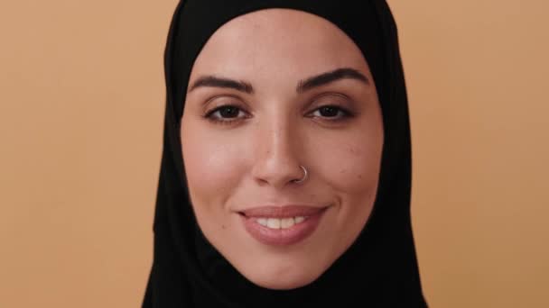 黒いヒジャーブを着た幸せなアラビア人女性のクローズアップは 彼女の目を開いて スタジオのベージュの壁の上に隔離されたカメラの前で笑っています — ストック動画