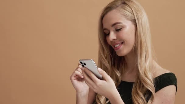 Nöjd Blond Kvinna Använder Sin Smartphone Stående Isolerad Över Beige — Stockvideo