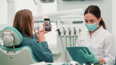 Koruyucu maske takan bir dişçi kadın, müşterisi klinikteki bir klinikte oturup selfie çekerken not alıyor.