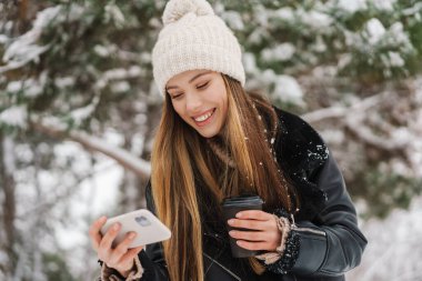 Mutlu kız, kış ormanlarında yürürken cep telefonu kullanıyor ve kahve içiyor.