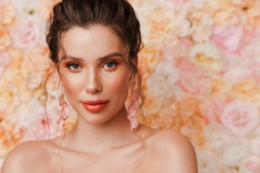 Küpe takan yarı çıplak genç bir kadın çiçek arkaplanından izole edilmiş kameraya bakıyor.