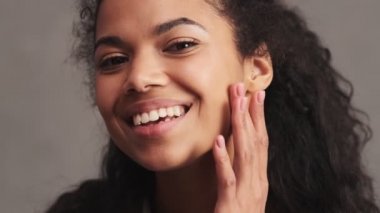 Gülümseyen Afrikalı Amerikalı bir kadının yakından çekilmiş görüntüsü stüdyodaki gri duvarın üzerinde duran tenine bakıyor.