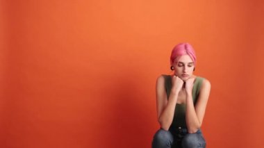 Pembe saçlı genç bir kadın yan tarafa bakıyor. Stüdyodaki turuncu duvarın üzerinde tek başına duruyor.