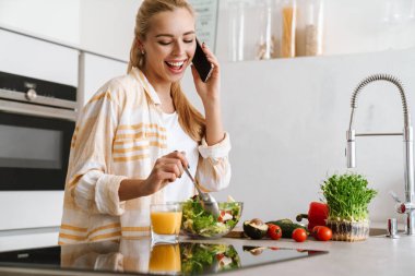 Mutfakta sağlıklı salata yaparken cep telefonu kullanan mutlu ve çekici bir kadının portresi.