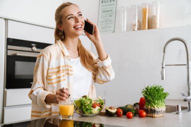 Mutfakta sağlıklı salata yaparken cep telefonu kullanan mutlu ve çekici bir kadının portresi.