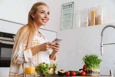 Mutfakta sağlıklı salata yaparken cep telefonu kullanan mutlu ve çekici bir kadının portresi.