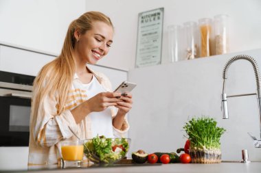 Mutfakta sağlıklı salata yaparken cep telefonu kullanan mutlu ve çekici bir kadının portresi.