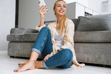 Evde otururken gülümseyen ve cep telefonu kullanan mutlu sarışın kadın.