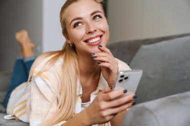 Mutlu sarışın kadın gülümsüyor ve evde koltukta yatarken cep telefonu kullanıyor.