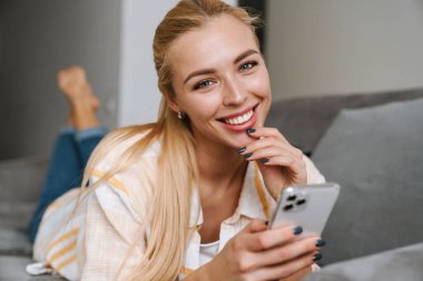 Mutlu sarışın kadın gülümsüyor ve evde koltukta yatarken cep telefonu kullanıyor.