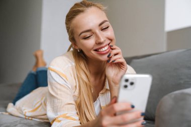 Mutlu sarışın kadın gülümsüyor ve evde koltukta yatarken cep telefonu kullanıyor.