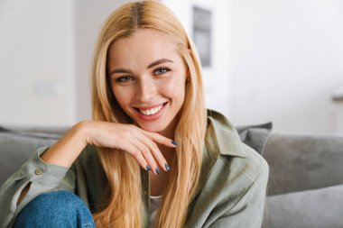 Evdeki koltukta otururken gülümseyen mutlu sarışın kadın.