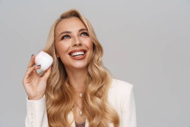 Gri arka planda izole edilmiş yüz kremiyle poz verirken gülümseyen mutlu muhteşem kadın.