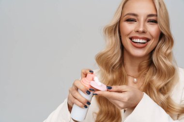 Gri arka planda izole edilmiş köpük ve kozmetik sünger kullanırken gülümseyen mutlu kadın.