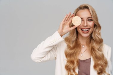 Gri arka planda kozmetik süngerle poz verirken gülümseyen mutlu muhteşem kadın.