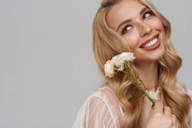 Güzel, gülümseyen, sarışın, elbiseli genç bir kadın. Mavi arka planda tek başına duruyor. Elinde güller var.