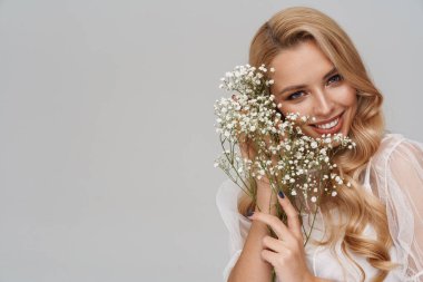 Gri arka planda saklı çiçeklerle poz verirken gülümseyen güzel mutlu kız.