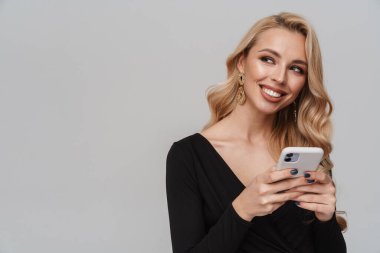 Gri arka planda izole edilmiş cep telefonu kullanırken gülümseyen lüks küpeli mutlu kız.