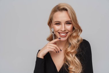 Gri arka planda izole edilmiş kameraya gülümseyen süslü küpeler takan mutlu sarışın kadın.