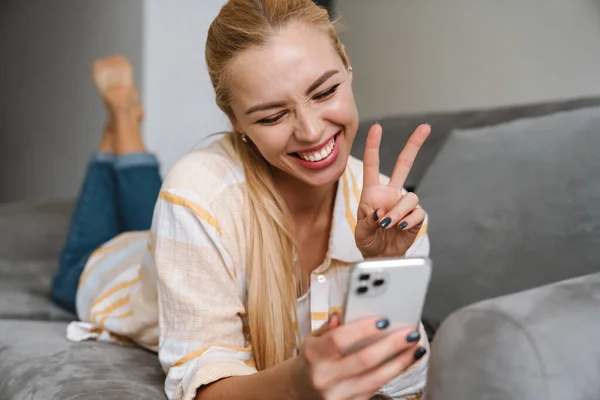 Щаслива Блондинка Показує Знак Миру Використовує Мобільний Телефон Дивані Вдома — стокове фото